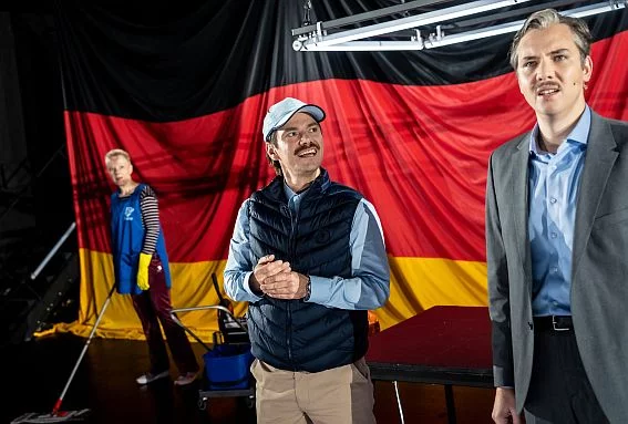 Drei Männer stehen vor einer Deutschlandflagge nebeneinander. Der Mittige grinst den Rechten an. | © Matthias Jung